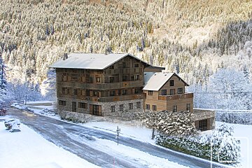 A multi-story wooden chalet with snow-covered roofs stands beside a snowy road, backed by a dense, snow-covered forest.