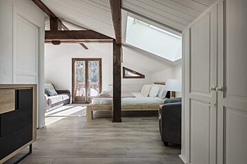 Modern attic bedroom featuring exposed dark wood beams, a skylight, a cozy bed, and a balcony in the background.