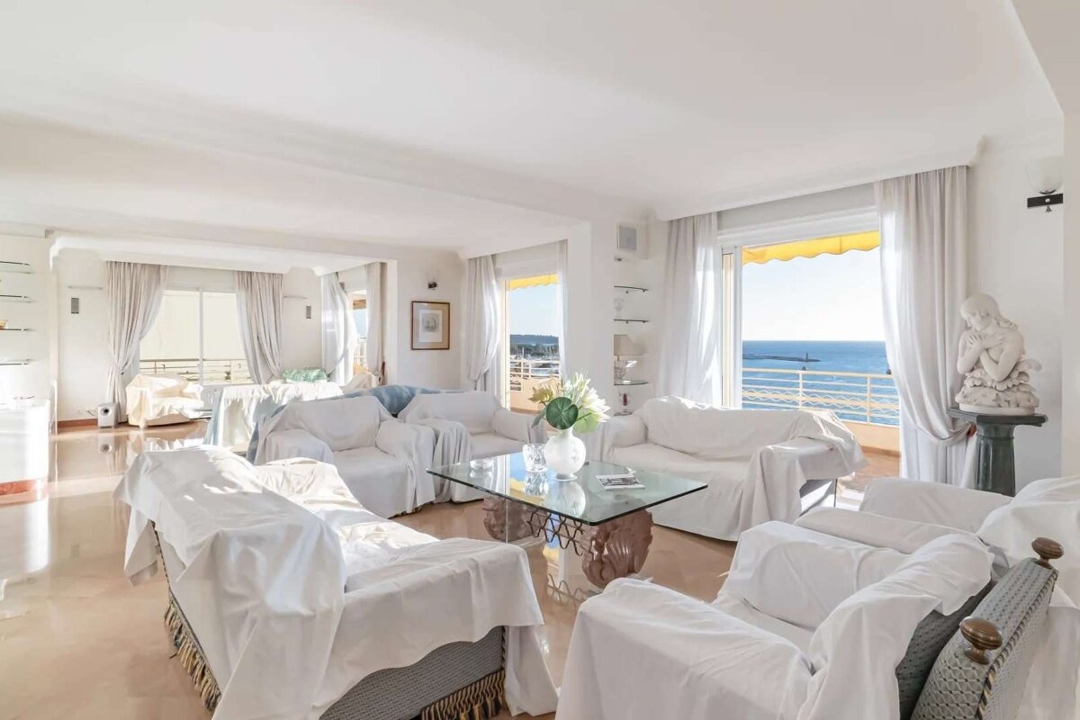 Bright, spacious living room with white slip-covered furniture, a glass coffee table, and an expansive ocean view from large windows.