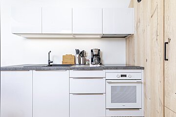 A minimalist kitchen with white cabinets, grey countertops, a built-in oven, a coffee maker, and a light wood wall panel.