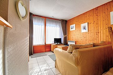 A cozy living room with a beige sofa, wood-paneled walls, tiled floor, and a bright window with sheer curtains.