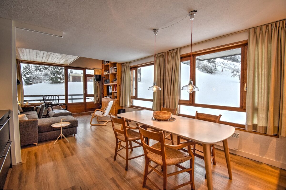 Cozy living/dining room with large windows overlooking a snowy landscape. Features a sofa, dining table, and warm wooden accents.
