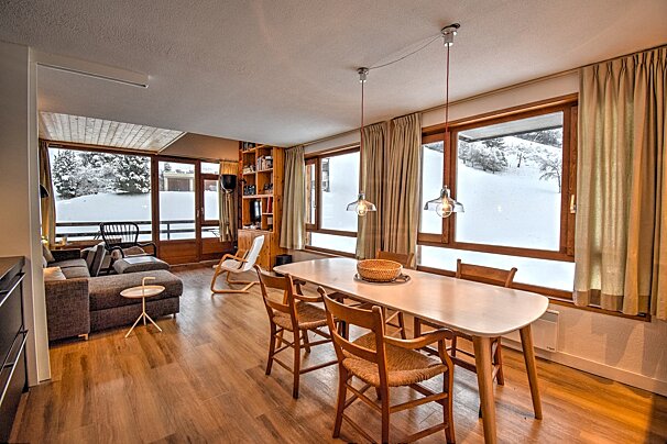 Cozy chalet interior with a dining table, living area, and large windows looking out onto a snowy hill.