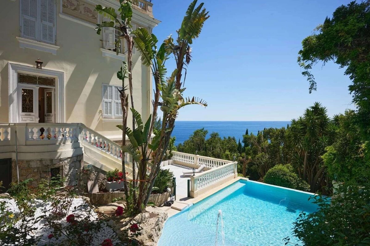 A luxurious villa with a sparkling pool and a terrace, offering panoramic views of the blue ocean, surrounded by vibrant green foliage.