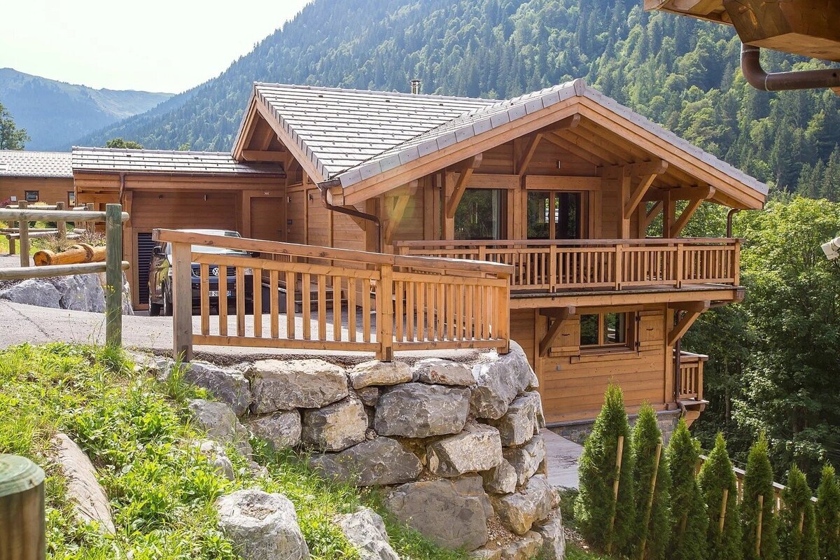 A beautiful wooden chalet with a balcony, supported by a stone wall, against a lush green mountain backdrop on a sunny day.