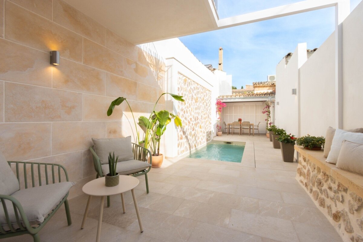 A serene outdoor courtyard features a small pool, modern green chairs, a stone-faced bench with cushions, and lush plants under a sunny sky.