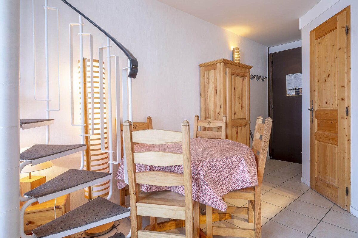 A dining area featuring a white spiral staircase, a round wooden table with a red patterned tablecloth, four wooden chairs, and a wooden cabinet.