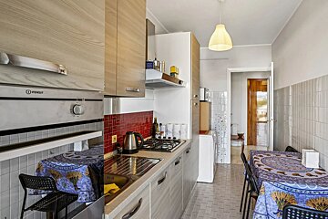 A narrow, modern kitchen with wooden cabinets, a sink, stove, and a small dining table draped in a blue tablecloth.