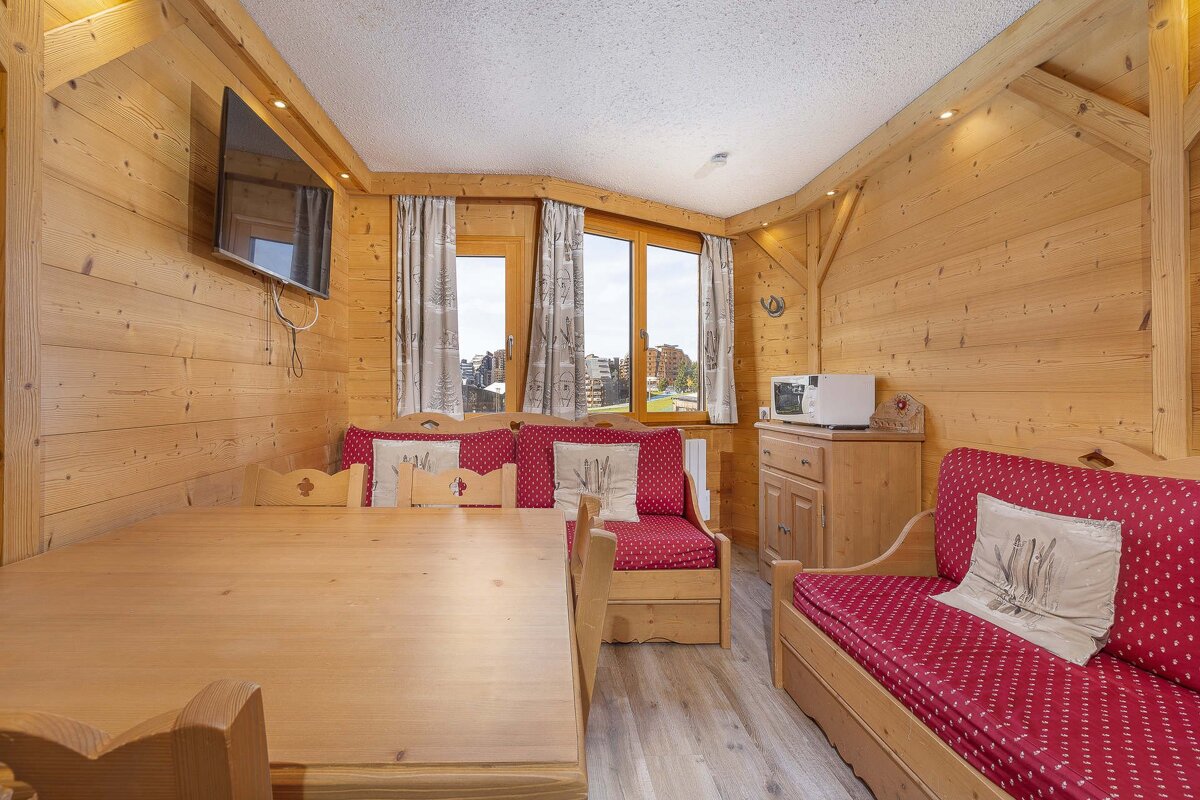 Rustic wood-paneled chalet interior with a dining table, red cushioned bench seating, a TV, and a window overlooking a village.