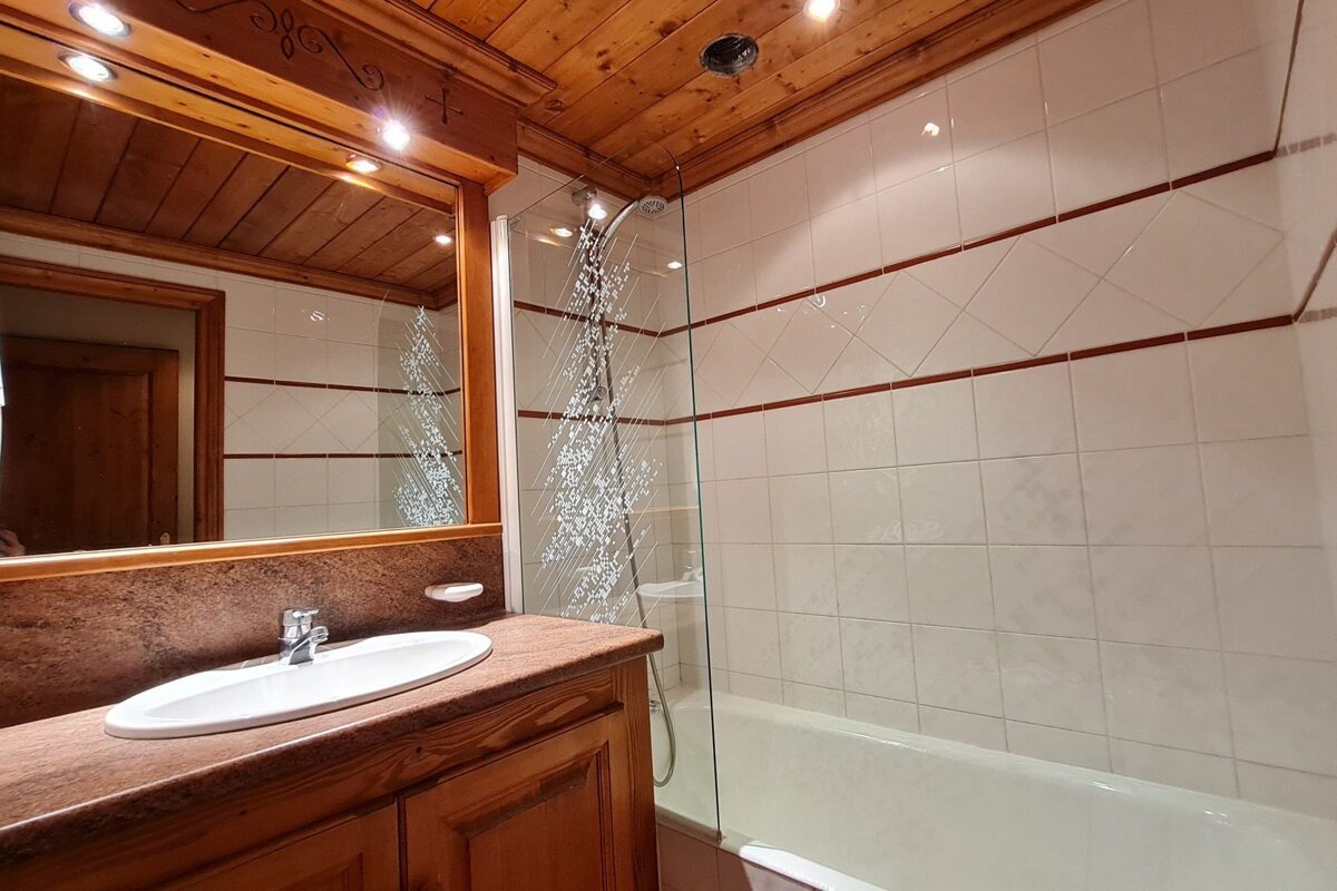 A cozy bathroom featuring a wooden ceiling and vanity, white tiled walls with brown accents, a sink with a mirror, and a bathtub with a glass shower screen.
