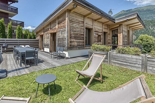 A rustic wooden chalet with a large deck, outdoor dining table, and lawn chairs on a sunny day in the mountains.