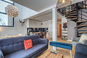 A modern, open-plan living room with a blue sofa, dining area, and a sleek kitchen. A black spiral staircase adds a chic touch to this contemporary space.
