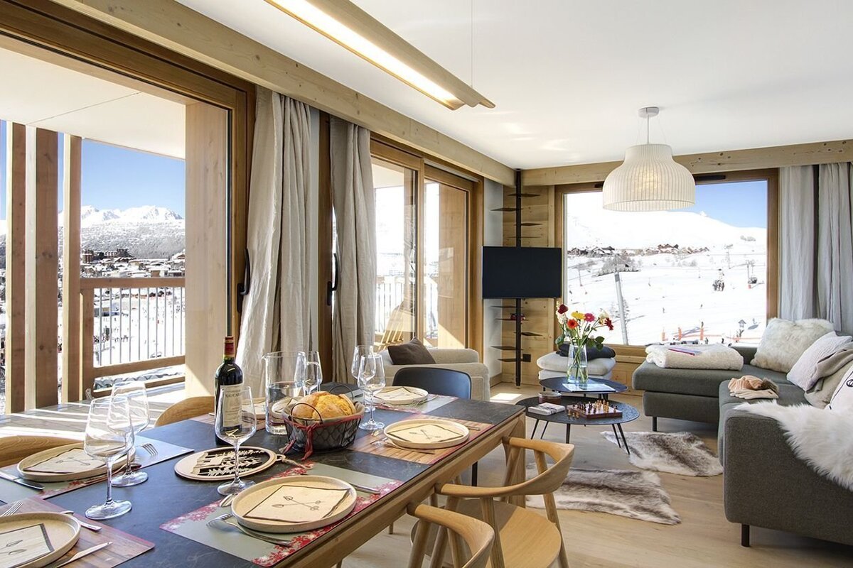 A stylish ski chalet interior with a dining table and living area, offering panoramic views of snow-covered mountains and ski slopes.