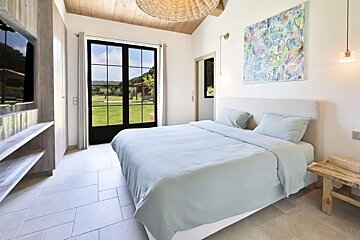 A bright, modern bedroom with a large bed, light blue bedding, tiled floor, and black-framed French doors opening to a green landscape. Abstract art adorns the wall.