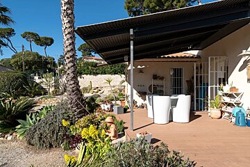 A sunny patio of a house with white outdoor chairs under an awning, flanked by a lush green garden and palm trees.