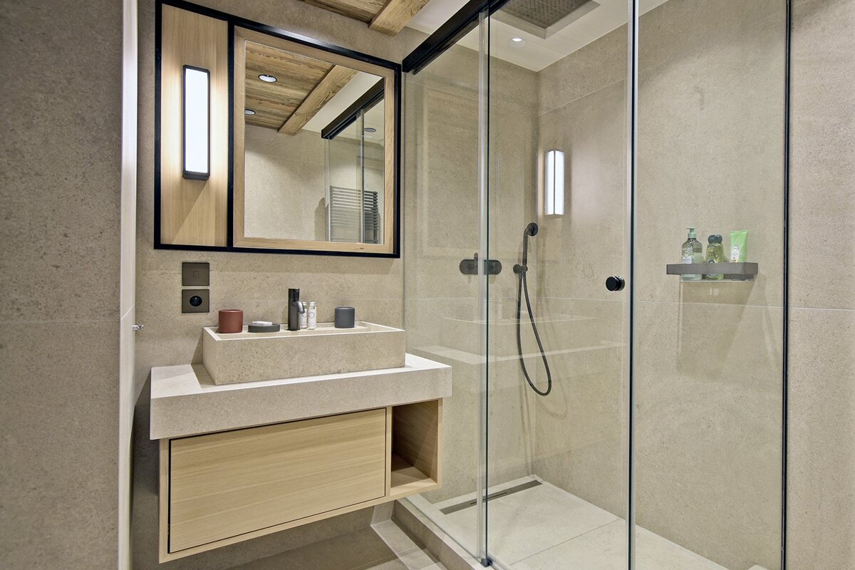 A modern, neutral-toned bathroom featuring a floating stone sink vanity, wood-framed mirror, and a sleek walk-in glass shower enclosure.