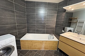 A modern bathroom with dark grey tiles, a wood-paneled bathtub, a washing machine, a wooden vanity with sink & mirror, and a towel warmer.