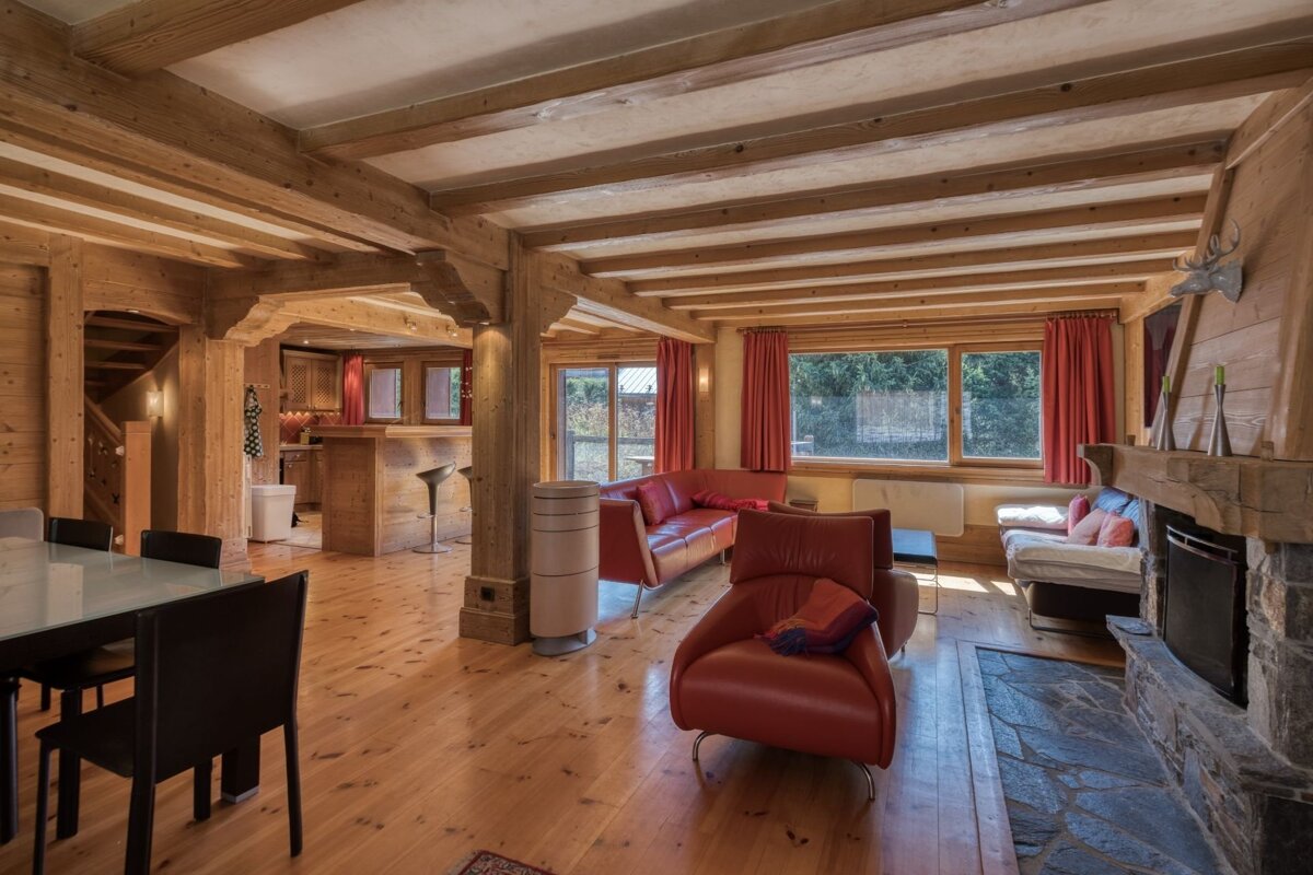 Warm, rustic wooden chalet interior with exposed ceiling beams, red leather armchairs, a stone fireplace, and a dining area.