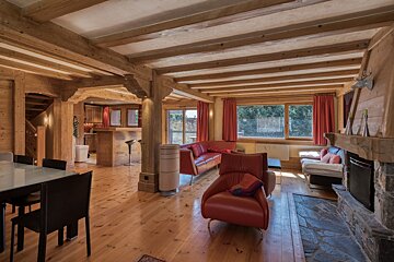 Cozy wooden chalet interior with exposed ceiling beams, red leather armchairs, a stone fireplace, and a dining table.