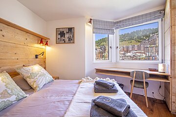 A cozy bedroom features a wooden headboard, folded towels on the bed, and a desk under a large window overlooking a scenic mountain town.