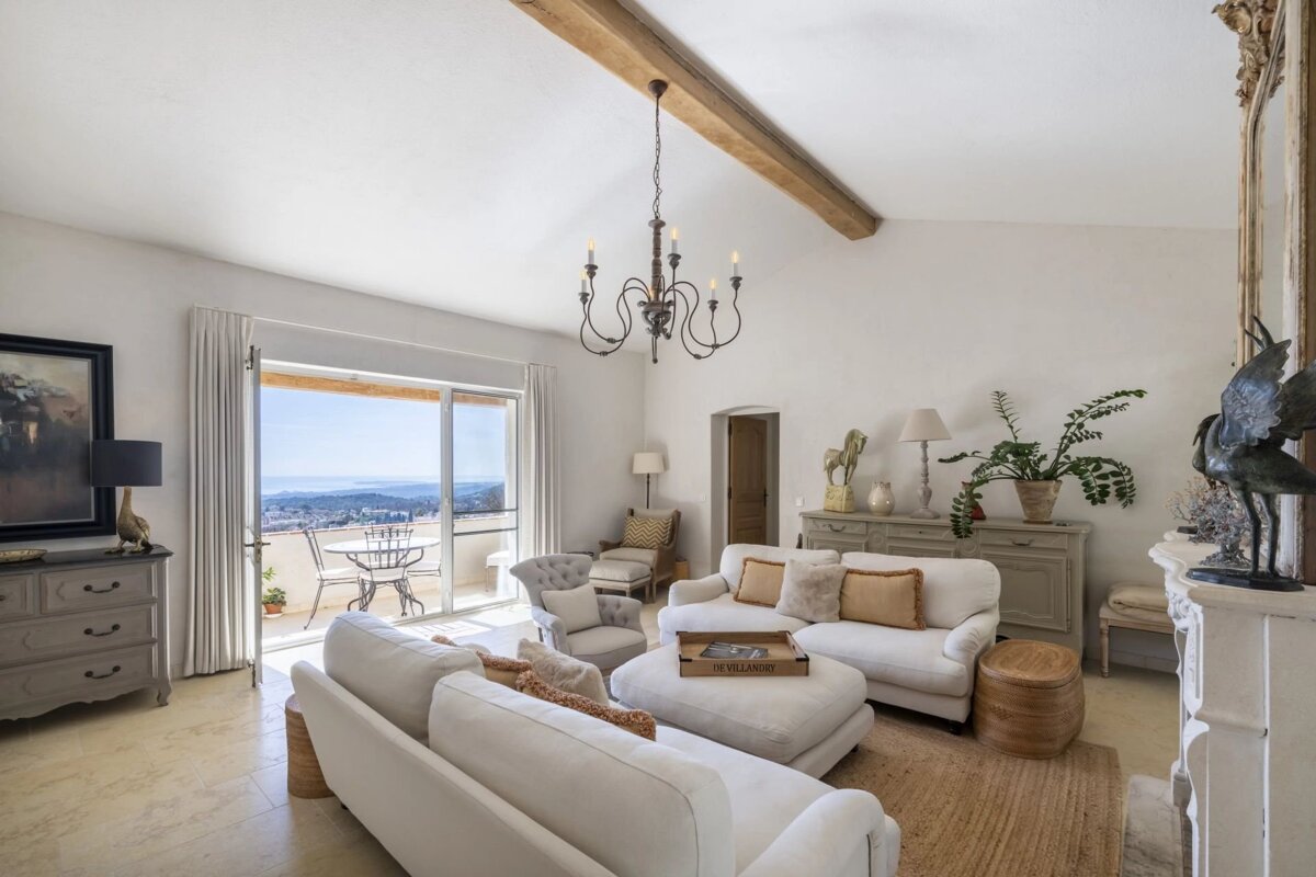 Bright, elegant living room with white sofas, neutral tones, a chandelier, wood beam, and a balcony offering scenic views.