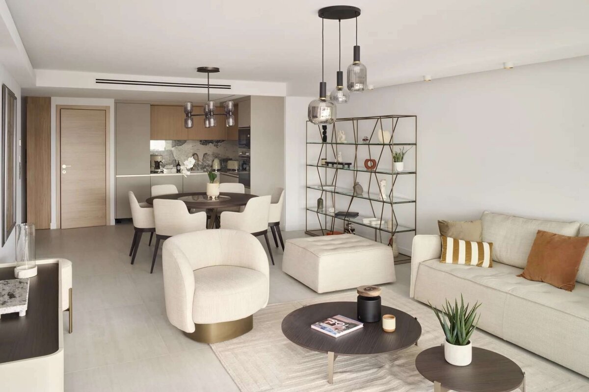 A modern, open-plan living room with a cream sofa, stylish armchairs, coffee tables, and a dining area, transitioning into a contemporary kitchen.