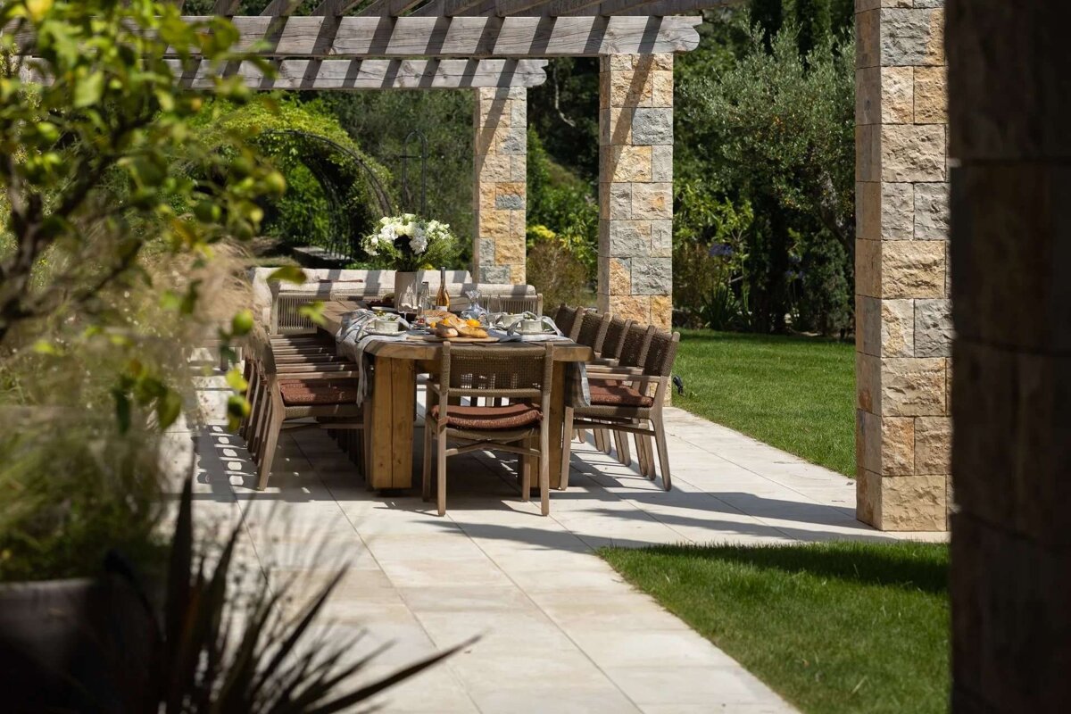 An inviting outdoor dining table under a wooden pergola, set for a meal in a lush garden with stone columns.