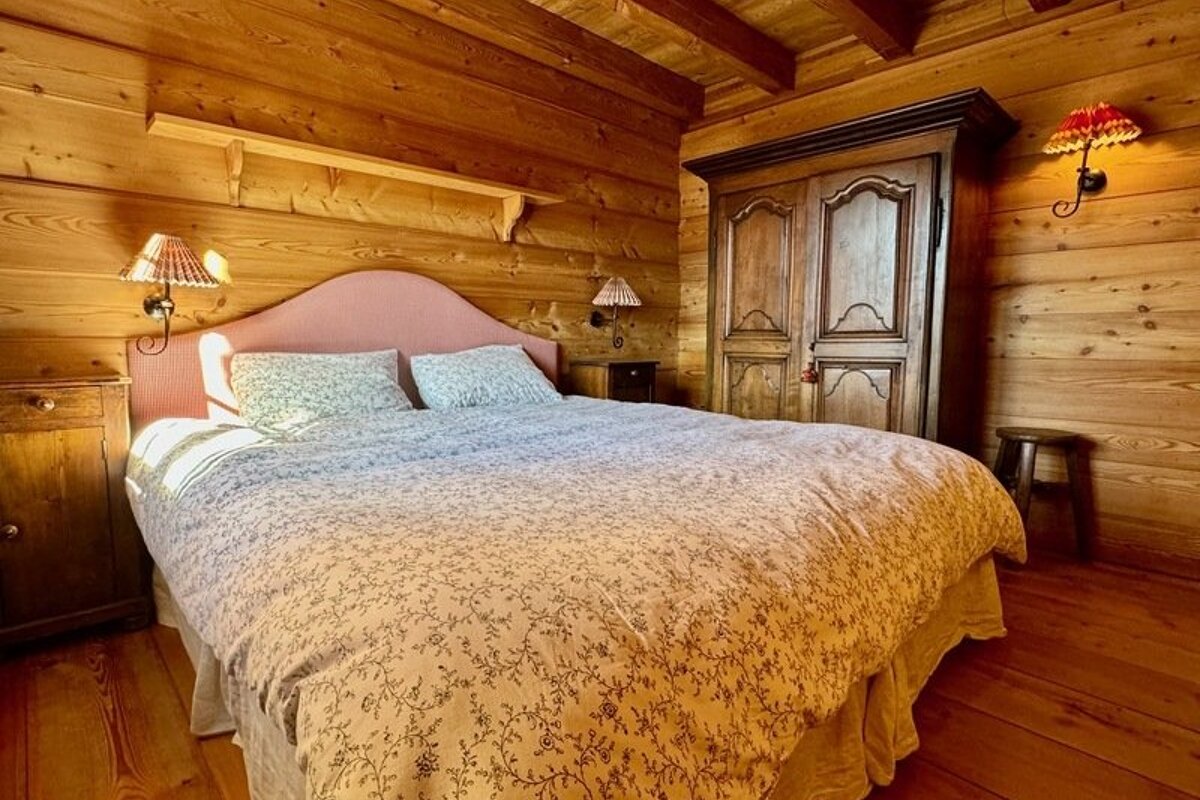 Cozy cabin bedroom with wood paneled walls, a large bed with a pink headboard, patterned duvet, and rustic furniture.
