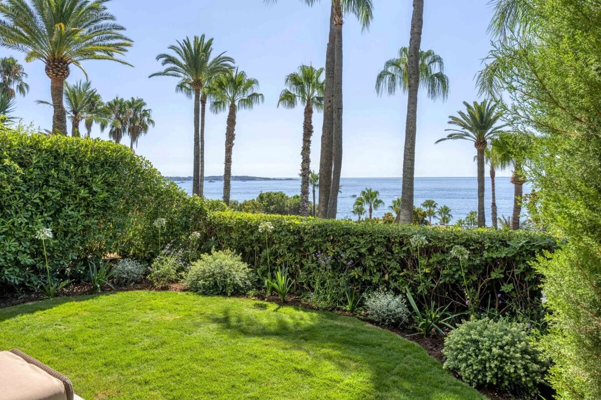 A lush garden with a green lawn and dense hedge, featuring tall palm trees against a bright blue sea under a clear sky.
