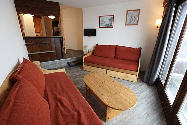 A rustic living room with two wooden sofas featuring orange cushions, a coffee table, wall-mounted TV, and a bar area with steps.