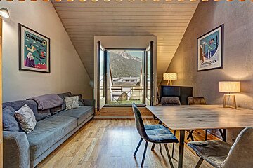 Cozy attic apartment featuring a grey sofa, dining table, Chamonix art posters, and window doors opening to mountain views.