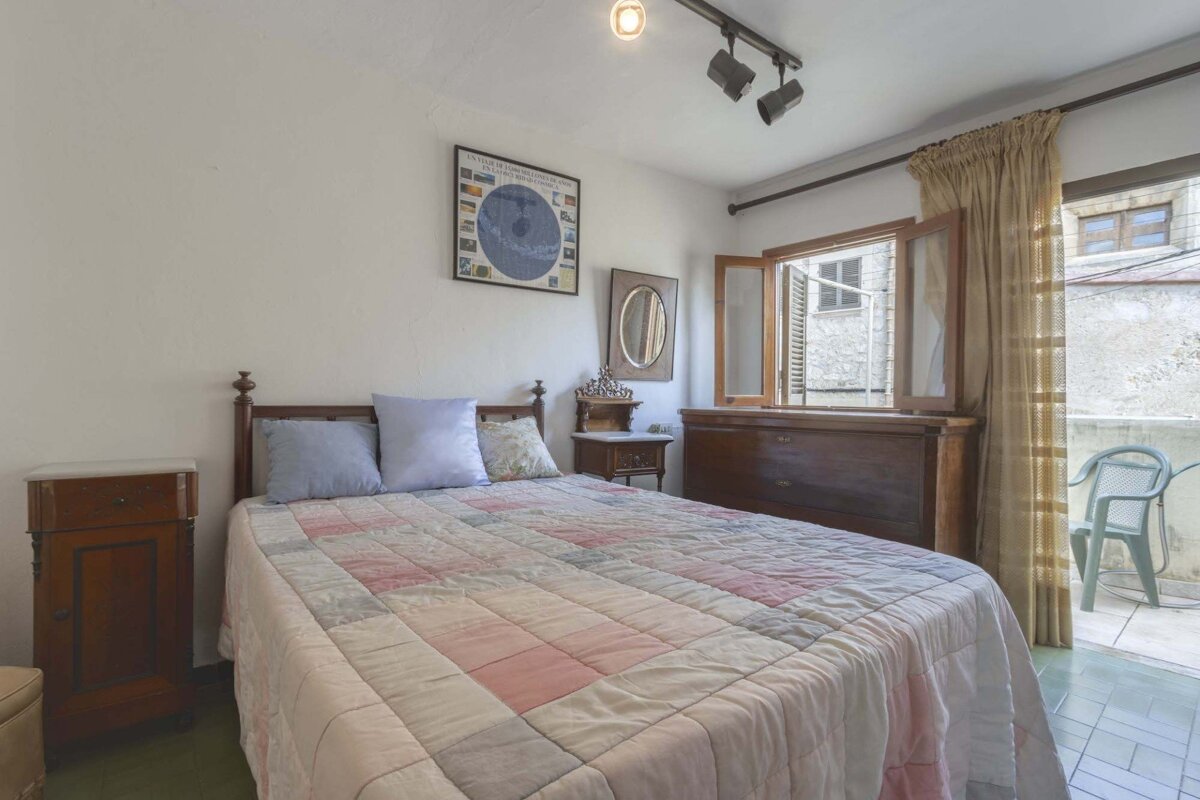 A cozy bedroom with a patchwork quilt on the bed, wooden furniture, an open window overlooking a street, and a small balcony with a chair.