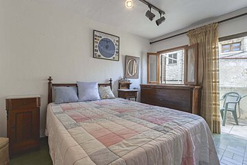 A cozy bedroom with a patchwork quilt on the bed, wooden furniture, an open window overlooking a street, and a small balcony with a chair.