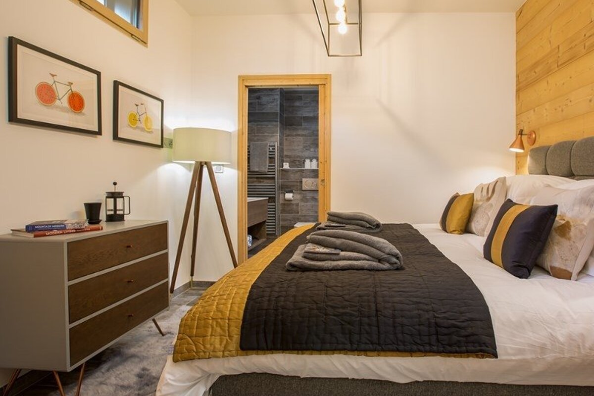 Cozy modern bedroom featuring a bed with grey and yellow accents, a dresser, a tripod floor lamp, and an open doorway to a bathroom.
