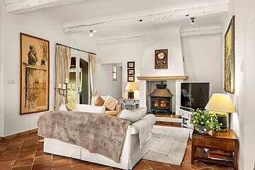 A cozy, rustic living room featuring a white sofa, a roaring wood stove, exposed ceiling beams, and warm terracotta tile floors.