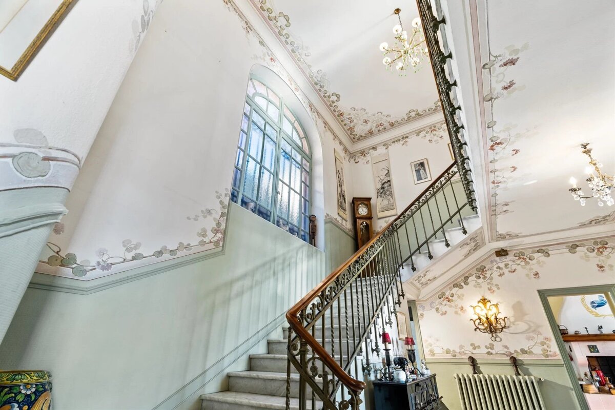 An elegant Art Nouveau staircase with delicate floral wall murals, an ornate railing, a grandfather clock, and classic light fixtures.