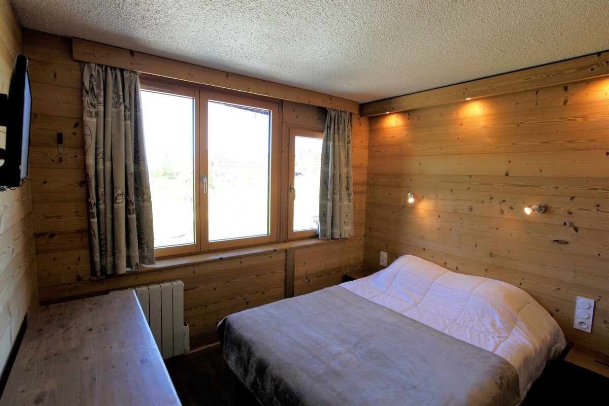 A cozy, wood-paneled bedroom featuring a large window, a neatly made bed, wall sconces, and a TV on the left.