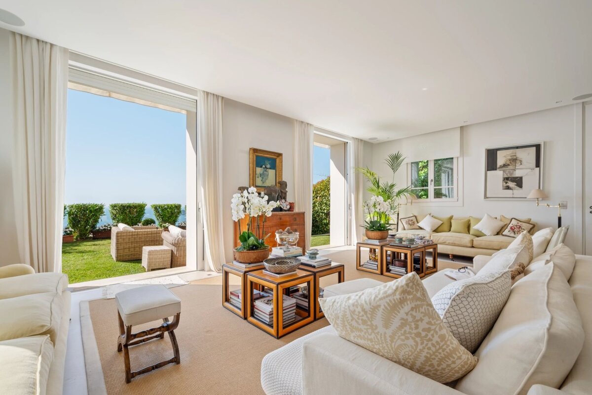A bright, elegant living room with plush white sofas, opening up to a sunny garden with beautiful sea views.