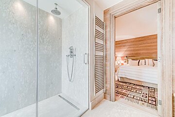 A modern walk-in shower with a rainfall head and towel warmer stands next to a doorway opening to a cozy bedroom with a wooden headboard and patterned rug.
