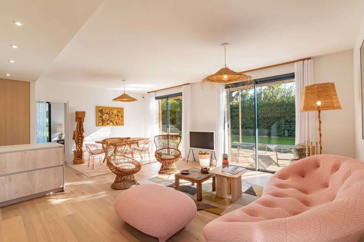 Sunlit modern living area with a pink quilted sofa, rattan chairs, light wood floors, and large glass doors opening to a lush green yard.