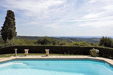 A scenic swimming pool offers a panoramic view of lush green hills, a distant blue sea, and a partly cloudy sky, with a tall tree and urns.