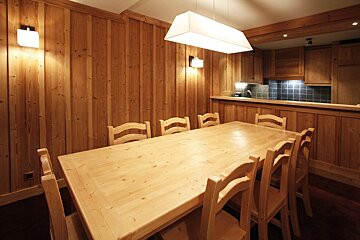 A warm, wood-paneled dining room features a long light-wood table with numerous chairs. A modern pendant light hangs above, with a kitchen visible in the background.