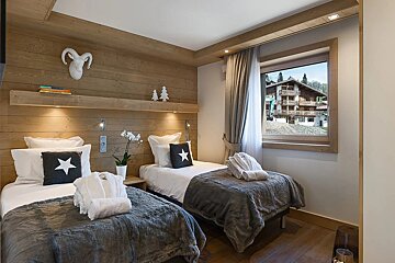A cozy, wood-paneled twin bedroom with two beds, fluffy robes, star pillows, and a window revealing a view of mountain homes.