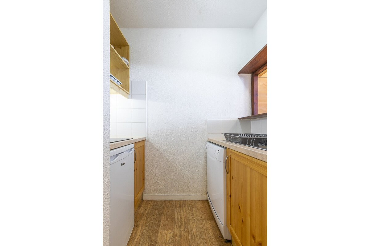 A compact galley kitchen with white walls, light wood cabinets, and appliances. Wooden floor and shelves complete the simple setup.