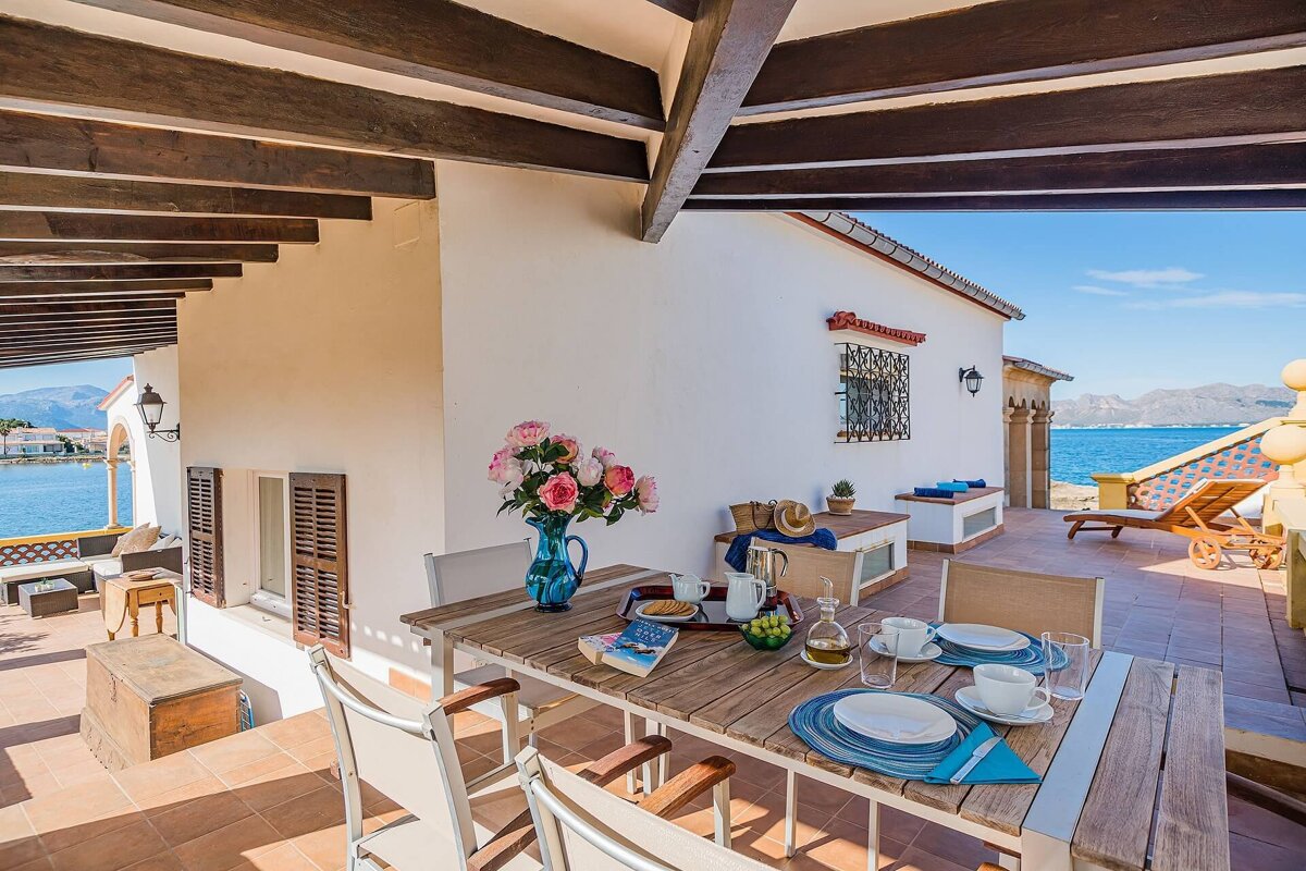 A Mediterranean terrace with a dining table, flowers, and a stunning view of the calm sea and distant mountains.
