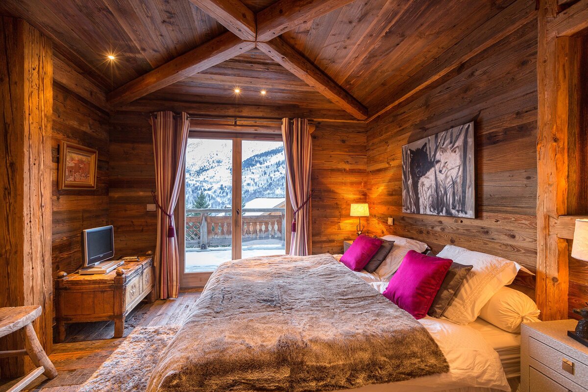 Cozy wooden chalet bedroom with a large bed, hot pink pillows, wolf art, and a beautiful snowy mountain view from the glass doors.
