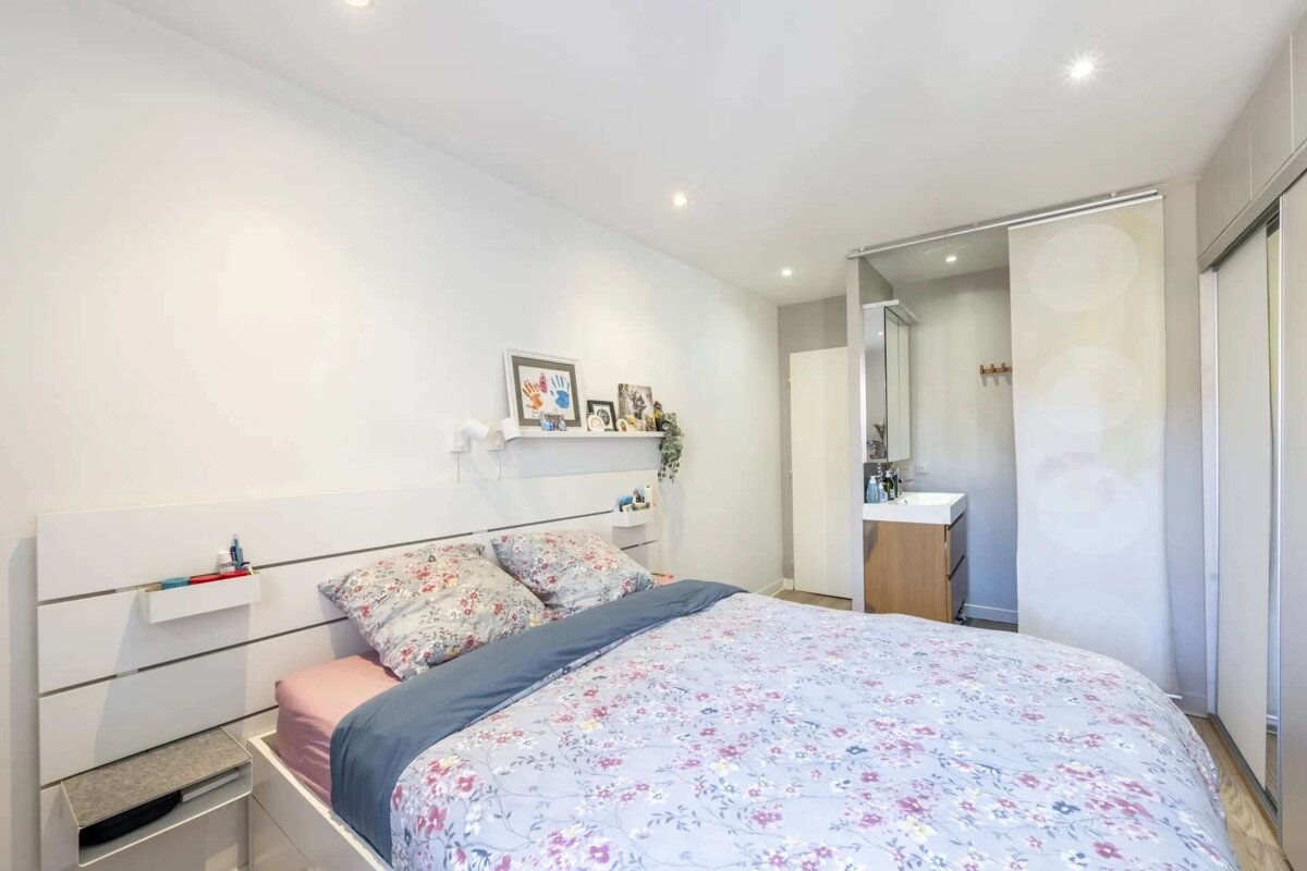 A bright bedroom with a floral bedspread, white headboard, and a visible en-suite bathroom featuring a sink and mirror behind a sliding screen.