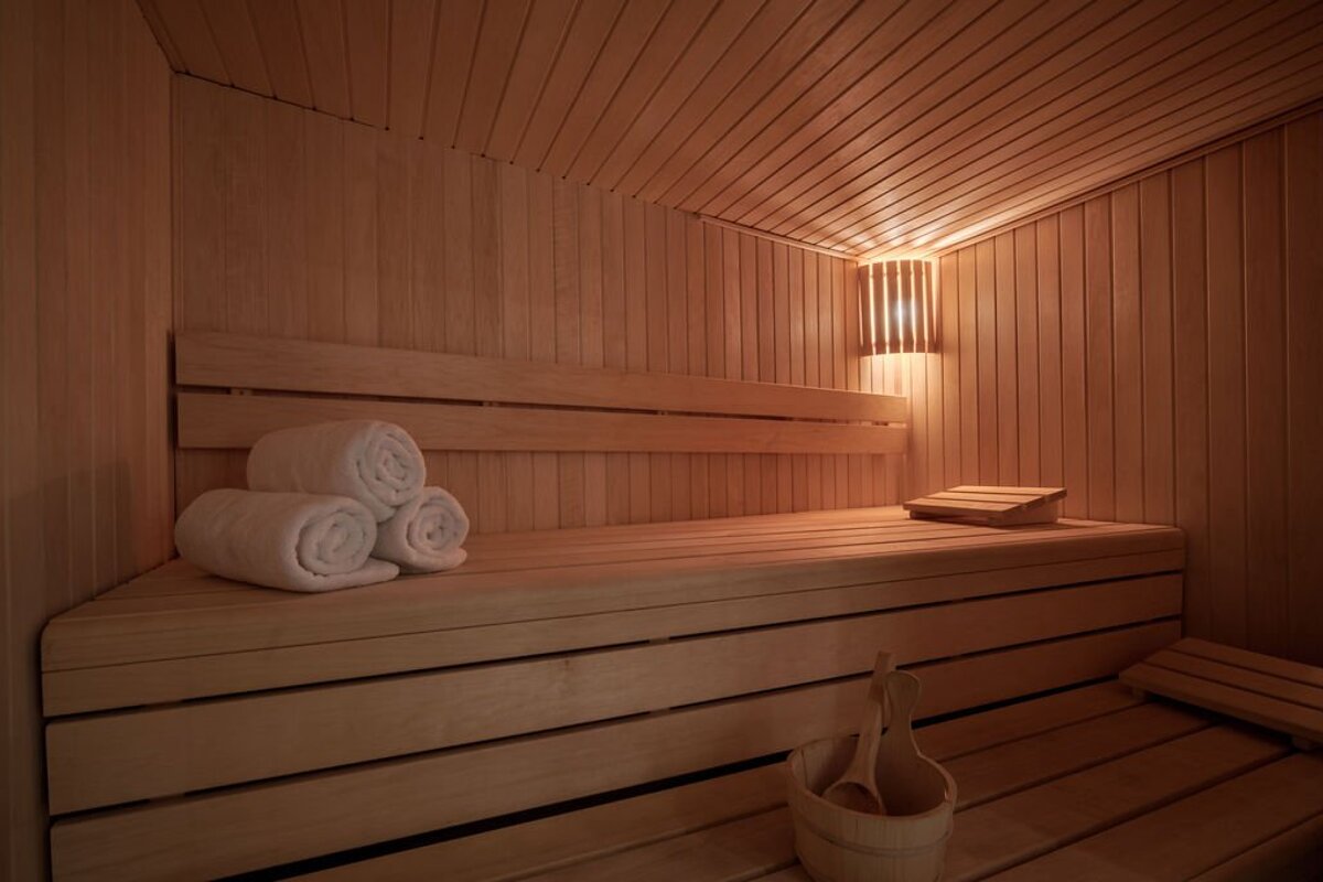A warm, inviting wooden sauna with soft lighting, featuring rolled white towels on a bench, a wooden headrest, and a bucket with a ladle.