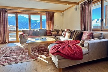 Cozy chalet living room with a large sectional sofa, red blanket, patterned rug, and large windows with scenic mountain views.