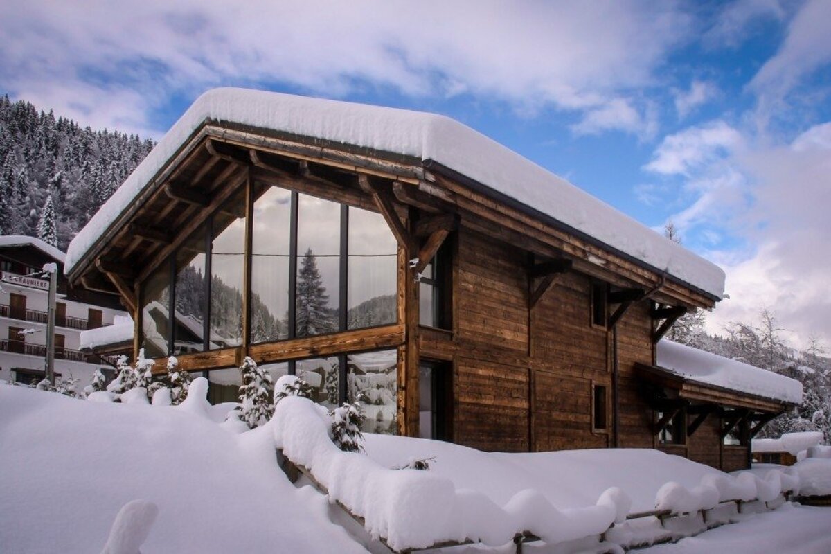 A modern wooden chalet covered in deep snow, featuring expansive windows reflecting a wintry mountain forest under a blue sky.